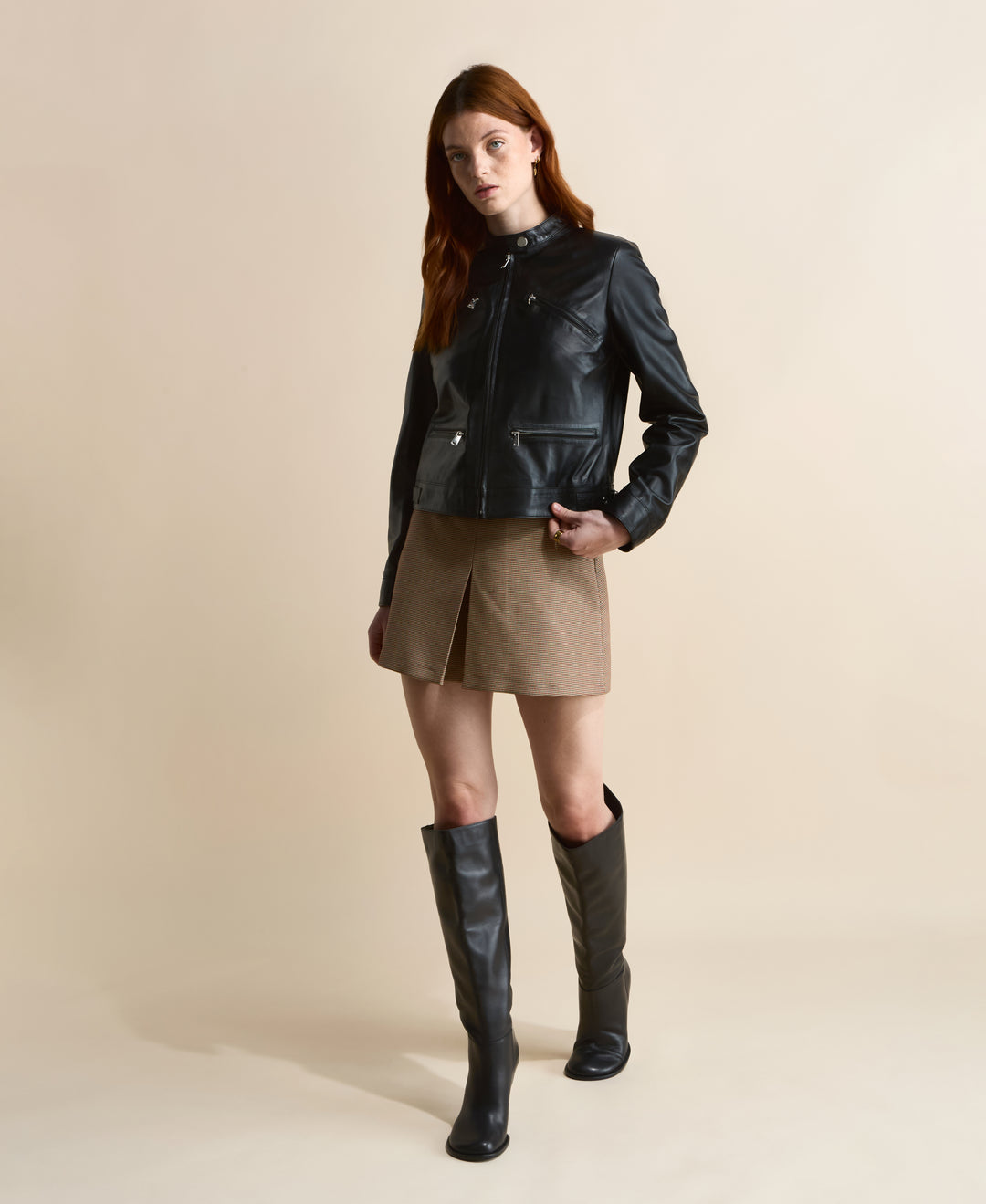 Black leather jacket with zip details styled with brown mini skirt and knee-high black boots, full body front view