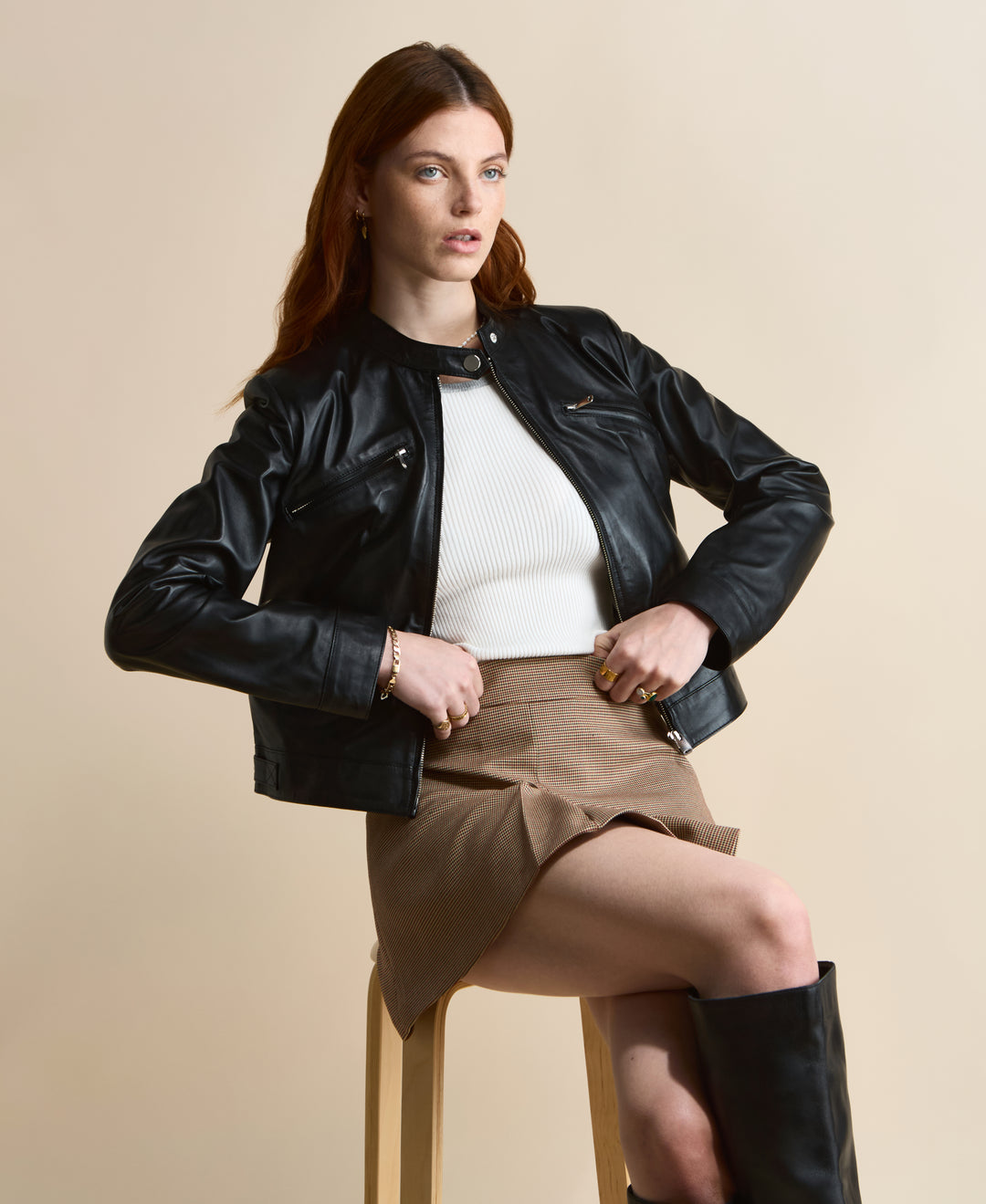 Black leather jacket styled with brown mini skirt and knee-high boots, seated pose