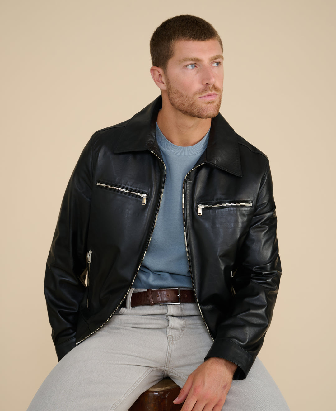 Men’s black leather jacket styled with light trousers seated pose