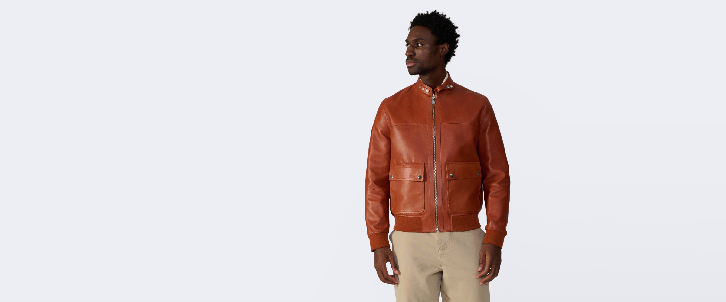 A man wearing a rust-orange leather bomber jacket and beige trousers, standing against a plain light gray studio background.