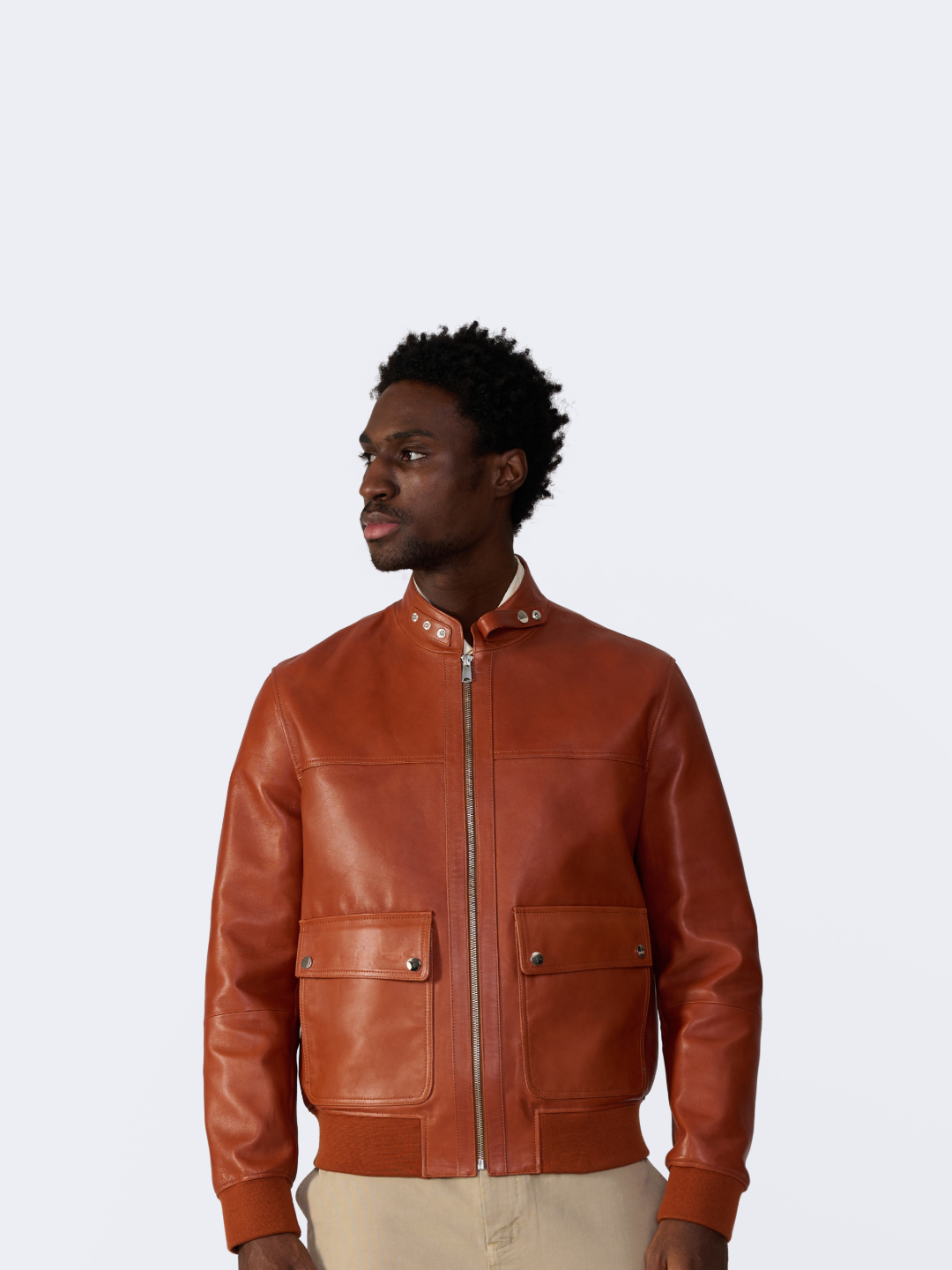 A man wearing a warm brown leather jacket with a silver front zipper, ribbed knit collar, and two front flap pockets, photographed against a plain light gray studio background.