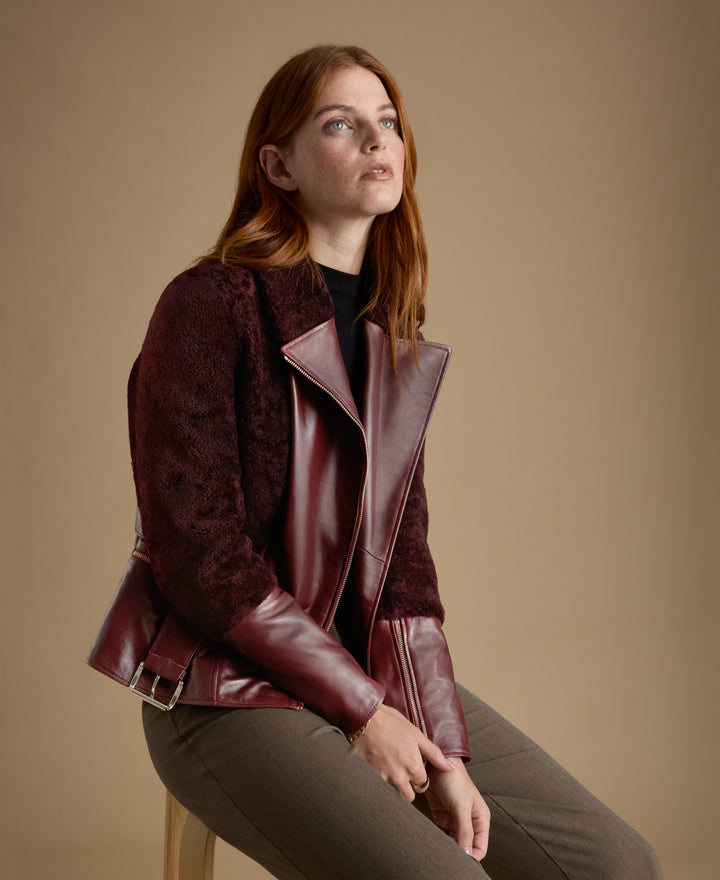Seated model wearing Alice burgundy leather and shearling jacket with tailored trousers