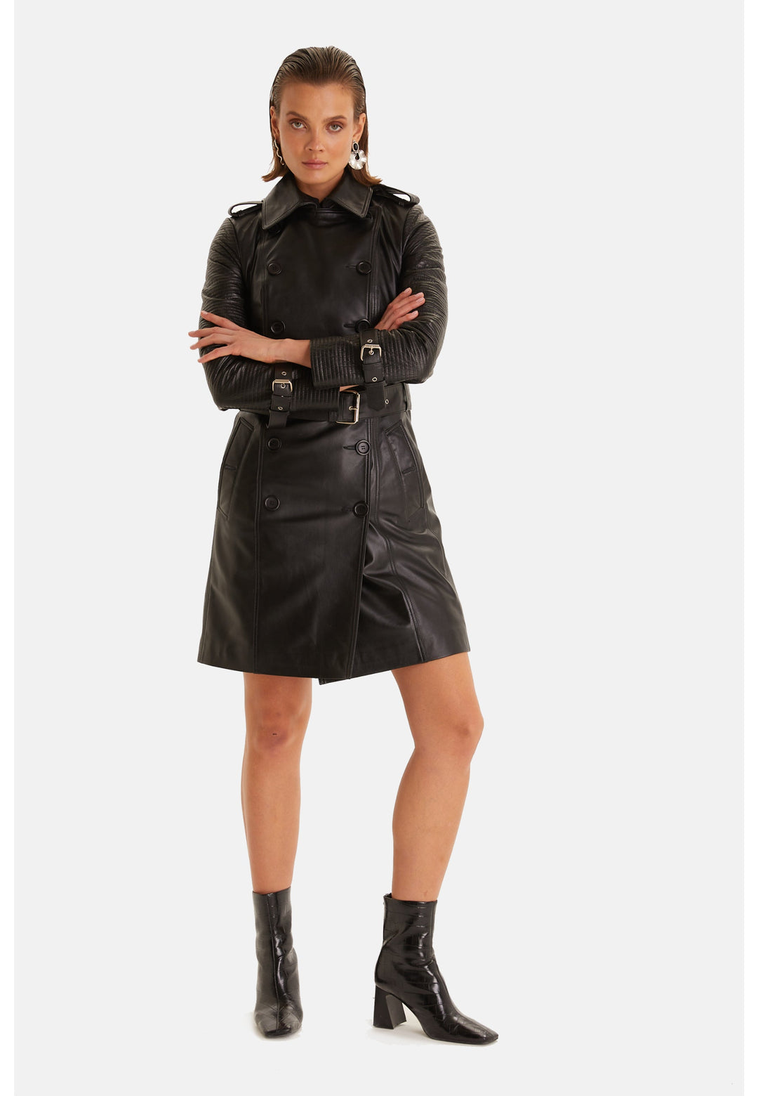 Model wearing the Greenwich black leather trench coat buttoned and belted, with arms crossed.
