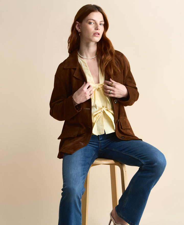 Seated model wearing the Vera chestnut brown suede jacket, showing the lapel collar and front flap pockets.
